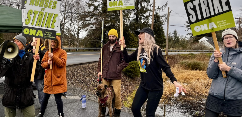 Six people, dressed for a chilly day, holding ONA "Nurses on Strike" picket signs, shout or sing against a gray Oregon sky alongside a road. One man holds the leash of a dog. Most people in the photo appear to be white women.