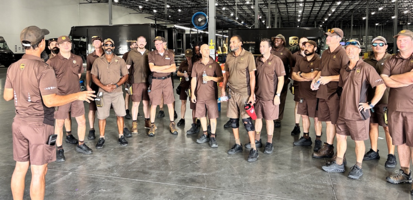 Fifteen workers in brown UPS uniforms stand in a semicircle while another uniformed worker speaks to them