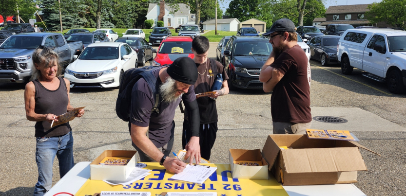 A group of Teamsters in a parking lot stand around a table with petitions and leaflets on it. One is filling out a sign-up sheet.
