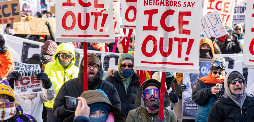 View of a section of a giant crowd marching, on a bright sunny day, bundled against extreme cold. Prominent picket signs that look screen-printed or stenciled say "Neighbors say ICE out!" in crisp red on white background.