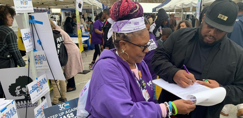 Outdoors at a public event, a woman in SEIU purple holds a clipboard while a man signs. The booth behind her is decked witht "SCHOOLS AND COMMUNITIES FIRST" signs and one that says "Step up against mass incarceration."