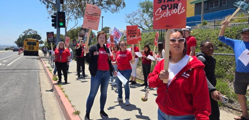 Teachers in red picket alongside a road with “Fully Staff Our Schools signs”