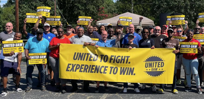 A large group stands behind a yellow banner saying “United to Fight, Experience to Win”