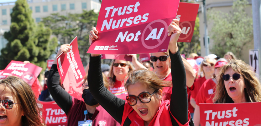 Women workers in red scrubs rally and chant, outdoors on a sunny day. They hold printed NNU signs. The woman most visible, in big sunglasses, holds a printed sign reading "Trust nurses, not A.I."