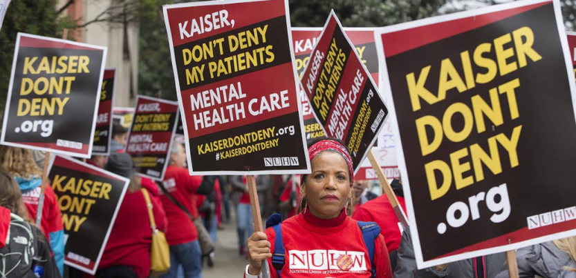 Strikers in red march with printed picket signs. In the center is a Black woman with a serious expression. Her sign reads "Kaiser, don't deny my patients mental health care. KaiserDontDeny.org."