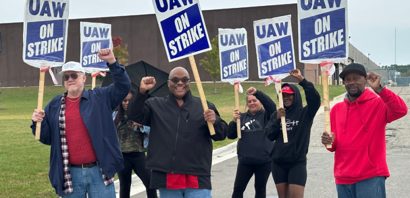 Six smiling workers carrying "UAW On Strike" picket signs raise their fists in the air outside a GM CCA facility. The workers are a mix of Black and white, men and women. Some of the picket signs are wrapped in plastic bags, and one worker carries an open umbrella.