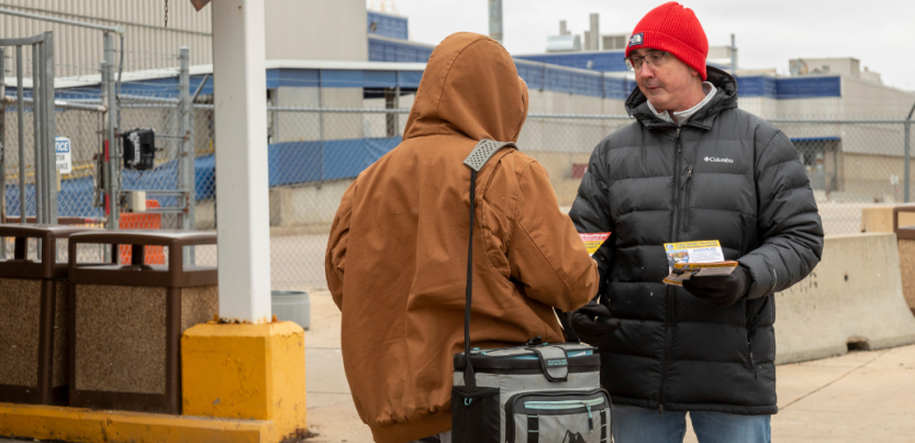 Outside an auto plant, Shawn Fain (right) in a heavy coat and red hat talks and hands a flyer to worker in a brown hooded coat whose back is to us.