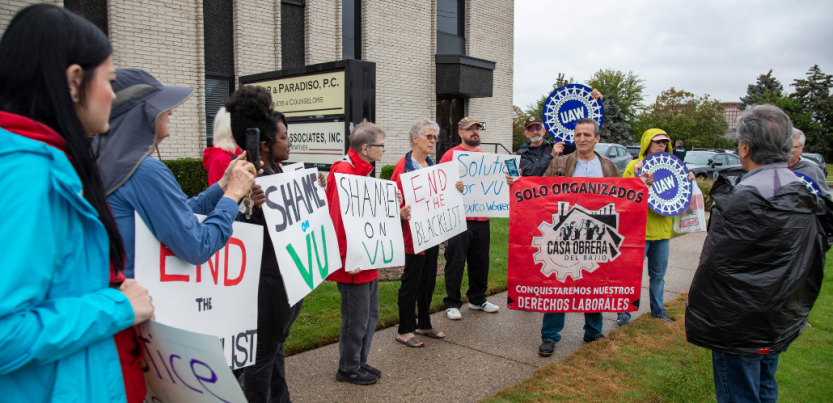 People with signs stand on grass outside an office building. The man speaking holds a red banner that says, "Solo organizados conquistaremos nuestros derechos laborales. Casa Bajia Obrera." Others listening hold printed blue UAW logo signs and hand-written signs that say "Shame on VU" or "End the blacklist." 