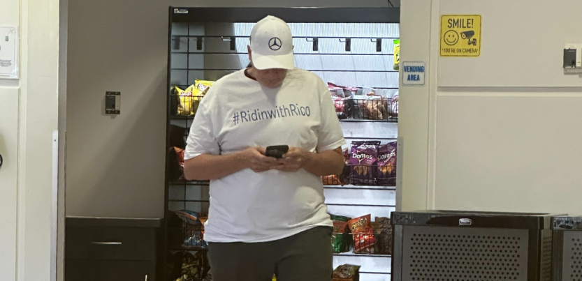 A person in a T-shirt with the message "#RidinwithRico" and a Mercedes logo hat looks down at their phone. They are standing in front of a vending machine.