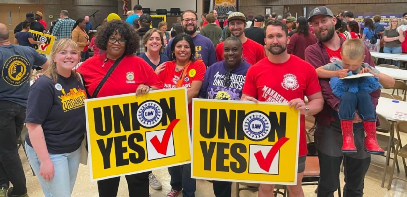 Ten smiling people pose together in a crowded meeting room. Two are holding huge yellow "Union Yes" signs with the UAW logo. Most are wearing red, blue, or purple T-shirts with UAW logo or organizing-themed slogans. The man on the far right holds a small child in his arms.