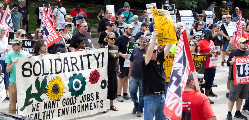 A rally with 30 or so people with a mixture of colorful signs, including Painters Union and a “We Want Good Jobs and Health Environments” banner