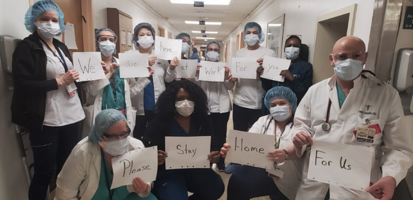 Masked health care workers in a hospital hold signs reading: "We... are... here... at... work... for.. you... Please... stay... home... for us."