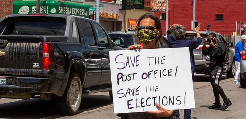 A masked protester holds a sign: "Save the Post Office! Save the Elections!" Behind the person, traffic and a "Coney Island" (diner) across the street
