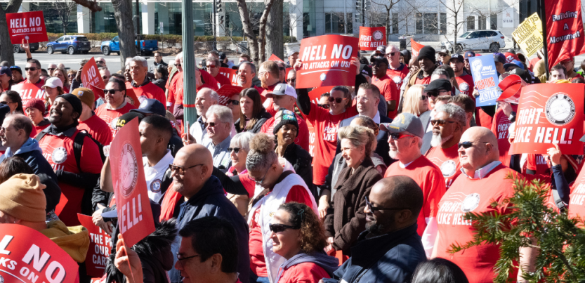 A large crowd of people in red "Fight Like Hell" shirts stand outside in bright sunlight. Some hold printed NALC signs with the slogans "Hell No to Attacks on Us" and "Fight Like Hell!"