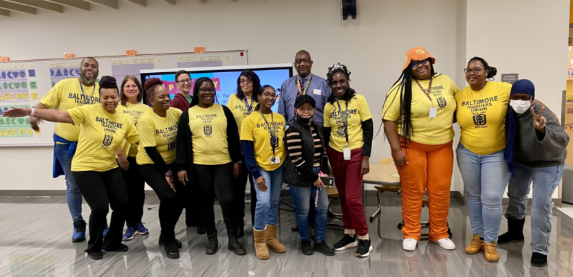 Fourteen workers, mostly Black and mostly women, pose smiling in a school classroom. Most are wearing yellow "Baltimore Teachers Union" T-shirts.
