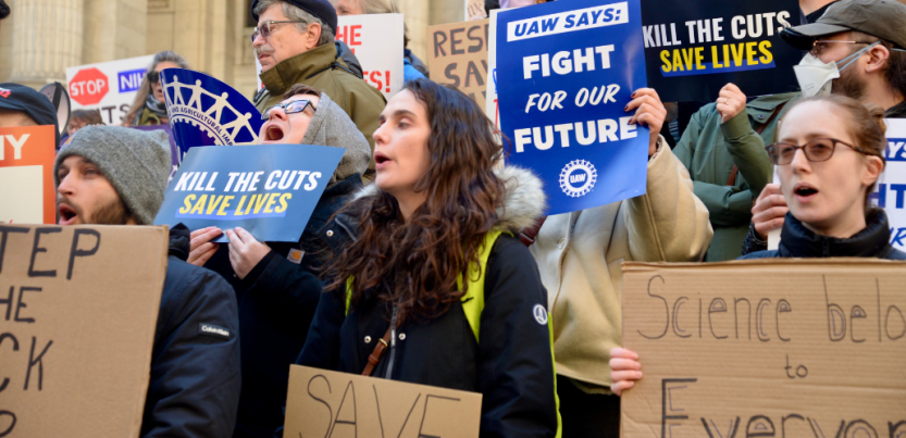 People, mostly white, rally outdoors on building steps. Printed signs say "Kill the cuts, save lives" and "UAW says: Fight for our future!" Handmade signs can't all be read but one says "Science belongs to everyone."
