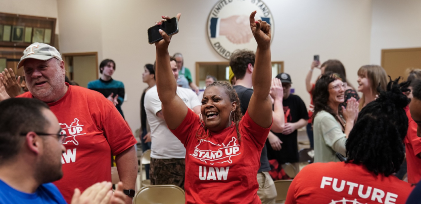 A Black woman in a red "Stand up UAW" T-shirt throws her hands in the air in celebration, phone in one hand, tears in her eyes. Other workers in matching shirts, and other people, stand nearby, all smiling, some clapping. In a room, looks like a union hall, with a logo on the wall that says "United we stand, divided we fall" around a handshake. 