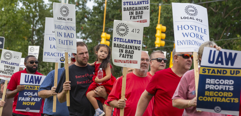 Auto workers march with signs that say “Stellantis Bargining in bad faith” One man is carrying a four-year old child.