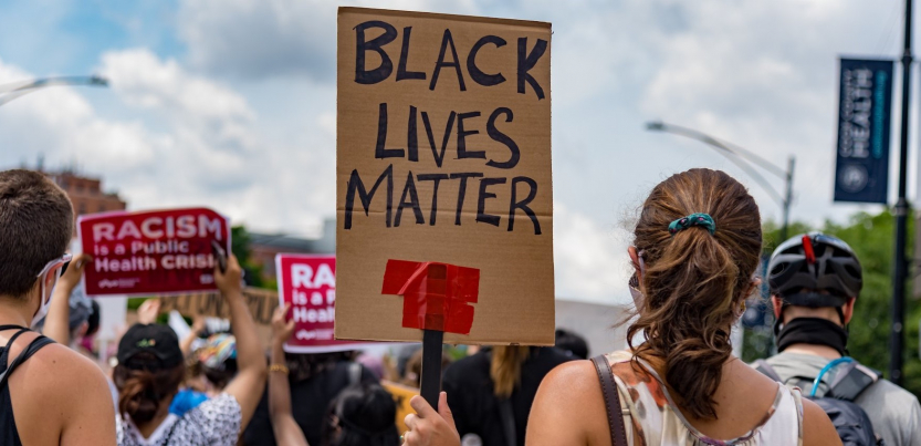 Crowd viewed from back. Sign in foreground, hand-lettered: "Black Lives Matter." Signs in background, printed: "Racism is a public health crisis" with the logo of National Nurses United
