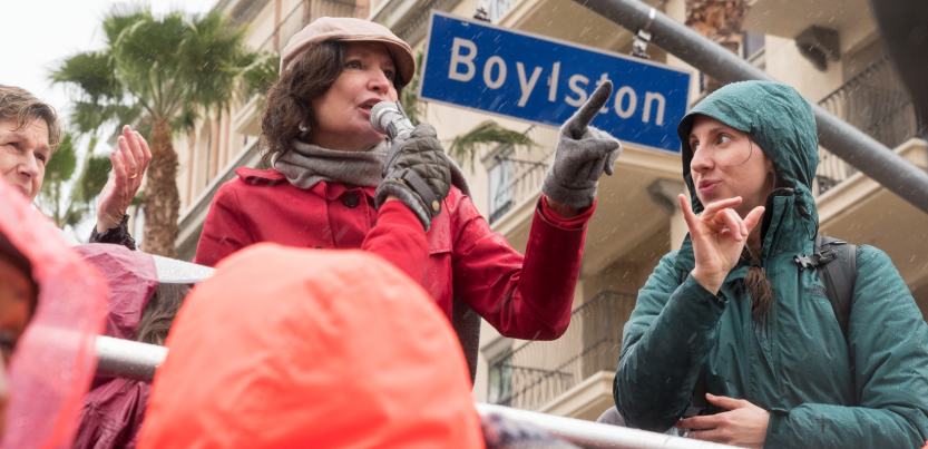 A woman speaks at a strike rally while another woman interprets in sign language. Behind them is a palm tree, an apartment buidling or hotel, and a street sign "Boylston"