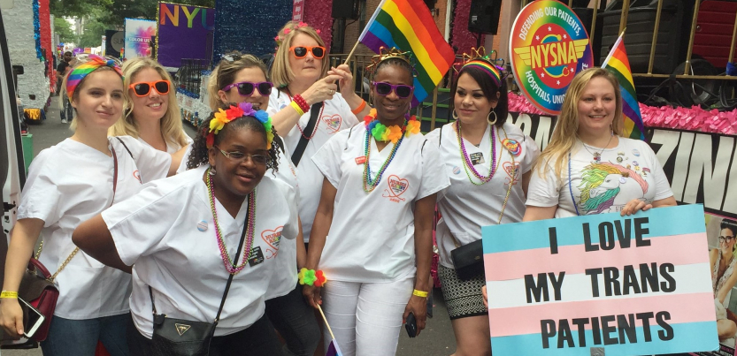 Nurses in white scrubs with rainbow flags and accessories; one carries a sign "I Love My Trans Patients" with the trans flag (pastel blue, pink, and white stripes)