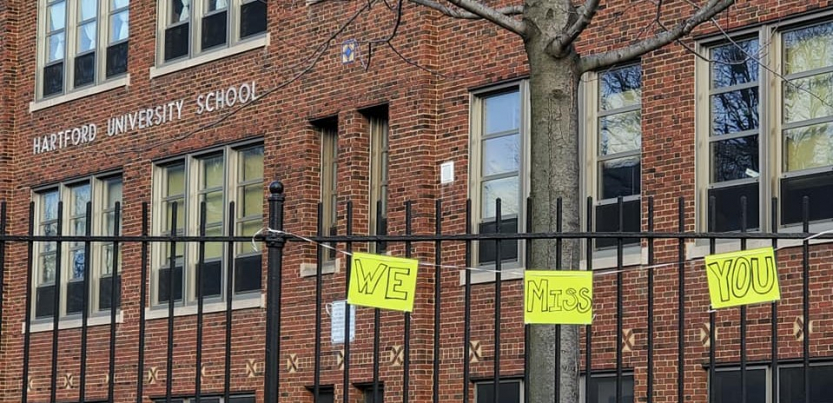 handwritten signs "we miss you" hang on the fence outside a brick building labeled Hartford University School