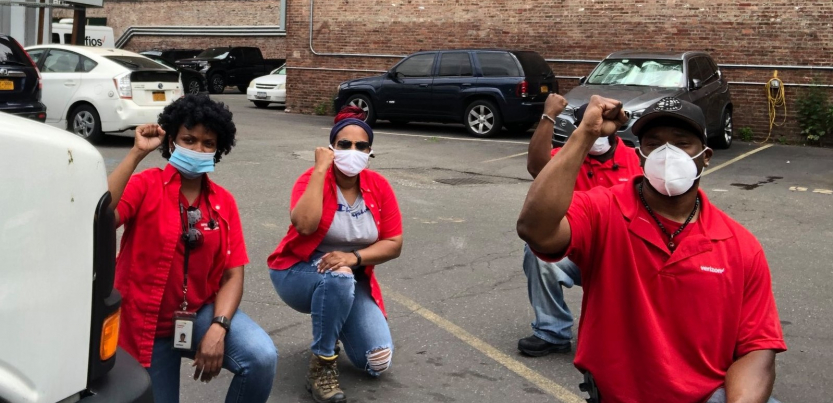 Four Black workers in red Verizon shirts kneel in a parking lot, fists raised.