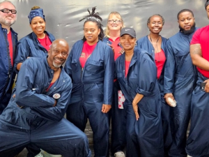 Nine workers (most of them Black, a mix of women and men) pose in red T-shirts and blue work overalls, apparently inside the plant. Their pose evinces camaraderie and confidence. One man in the front has a particularly dynamic pose with a lunge, crossed arms, and a big smile.