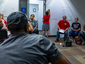 A group of labor activists sit together in a room while one writes ideas on a piece of flip chart paper on the wall.