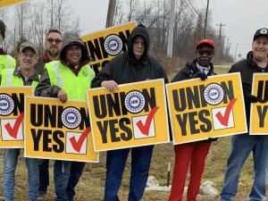 Seven workers, men and women, white and black, hold yellow “union yes” signs in front of the Lordstown water tower