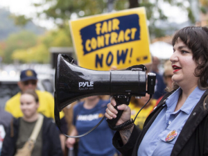 A woman with long dark hair speaks into a megaphone to a crowd that is visible beyond her, one holding a “Fair Contract Now” sign.