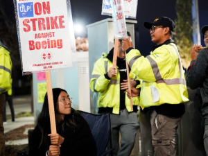 a young asian woman grips a sign that says on On Strike at Boeing. Others around her have signs. It’s nighttime.