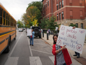 A teacher holds a sign between a school bus and the front of a school, with students visible in the background
