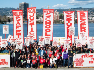 A crowd of hundreds is seen from afar and behind them there’s a body of water. They have vertical red on white banners saying “Health and Safety,” “Racial Justice,” “For the schools,” “That Students,” “Deserve.” 