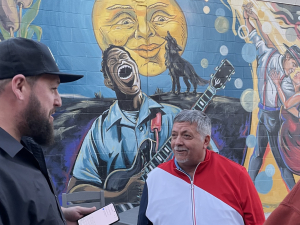 Three autoworkers talk while standing in front of a colorful mural with a painting of a man singing and playing guitar, a wolf howling, and a couple dancing the tango.