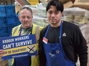 Two Kroger workers stand in front of pallets. One holds a sign that says "Kroger workers can't survive off of quarters."