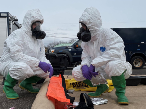 Two people in respirator masks and white hazardous materials suits squat and look at each other over a bright orange bag labeled "hazardous"