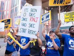 Nurses in blue T-shirts blow whistles and air horns outside a red-brick building with huge white columns.