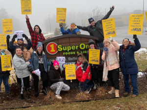 A group of 20 workers in winter clothes gather with yellow UFCW signs around a SuperOne Foods sign