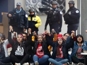 Teachers seated on sidewalk with joined hands raised; police standing behind them