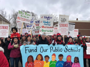 Strikers carry a banner "Fund Our Public Schools" with a rainbow of faces. Picket signs say "Strike" and "It's Time to Use Our Outside Voice." One kid is carried on an adult's shoulders.
