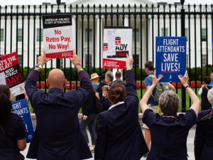 A line of seven flight attendants in uniform, viewed from behind, stands at the White House gates holding up various printed signs. Messages say: "AA makes billions, we can't pay rent." "Robert, what's the delay? We need a contract today!" "American Airlines flight attendants: No retro pay, no way!" "Flight attendants save lives!" "Corporate greed doesn't fly." "Trying to get pAAid." It appears the group includes men and women, and most are people of color.