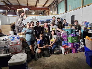 A group of fifteen workers pose inside a warehouse among boxes of diapers and other aid material