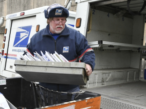 A man with a white beard in a blue postal uniform loads a tray of letters in to the back of a postal truck.