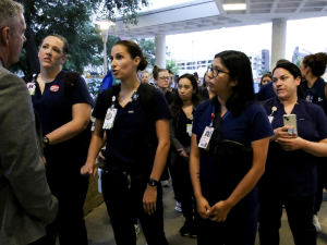 A group of nurses in dark blue scrubs with badges confront a man in a grey suit who is blocking their path.