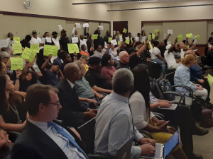 Inside a board meeting, dozens of union members and supporters hold up small yellow and white signs reading 'Fix Our Schedules.'