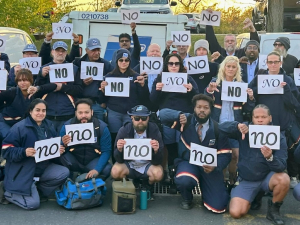 At least 32 letter carriers in uniform, holding "no" signs printed in various fonts, pose in a post office parking lot. The group looks diverse in race and gender. Several give the "thumbs-down" hand signal.