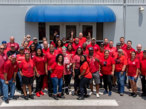 Forty workers in red t-shirts pose together outside a building with a blue awning.