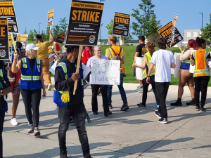 Workers (men and women, Black and white) picket outside in the sunshine. Some carry printed signs that say "Amazon workers on strike" with a Teamsters logo. One carries a hand-written sign that says "Stop the captive audience."