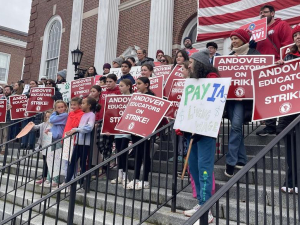 A large group of teachers stand in the steps of a large brick building holding red signs that say “Andover Educators on Strike” 